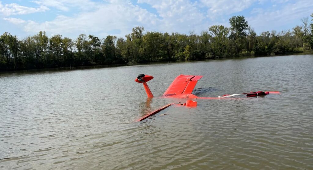 Tail section of a red Aviat S-1-11B aircraft protruding from a calm river during daylight, with the rest of the aircraft submerged. The tailwheel and horizontal stabilizers are visible above water.